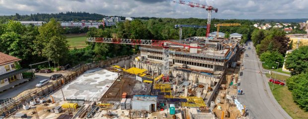 Ein Blick über die Baustelle nach Westen zeigt im Vordergrund das fertige Untergeschoss des Parkdeck- und Kantinengebäudes, dann der Tiefgaragen-Verbindungsbau (Schalarbeiten) und dahinter das künftige Hauptgebäude des Neubaukomplexes.